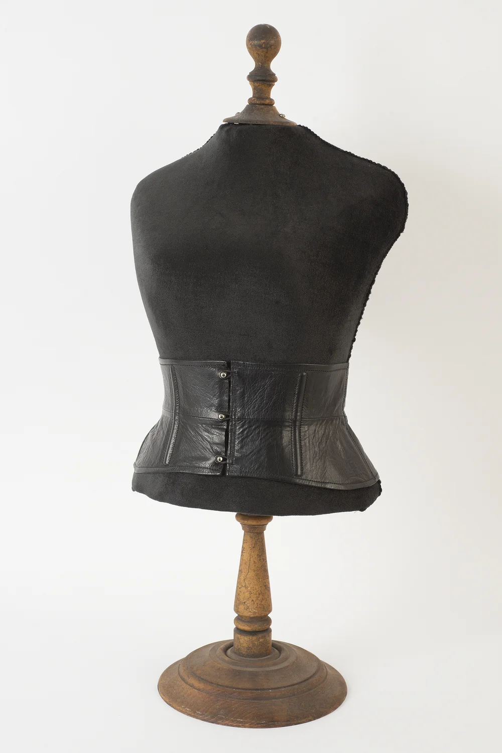 Antique mannequin torso wearing a black leather waist cincher with snap closures, dark wood stand, plain light background