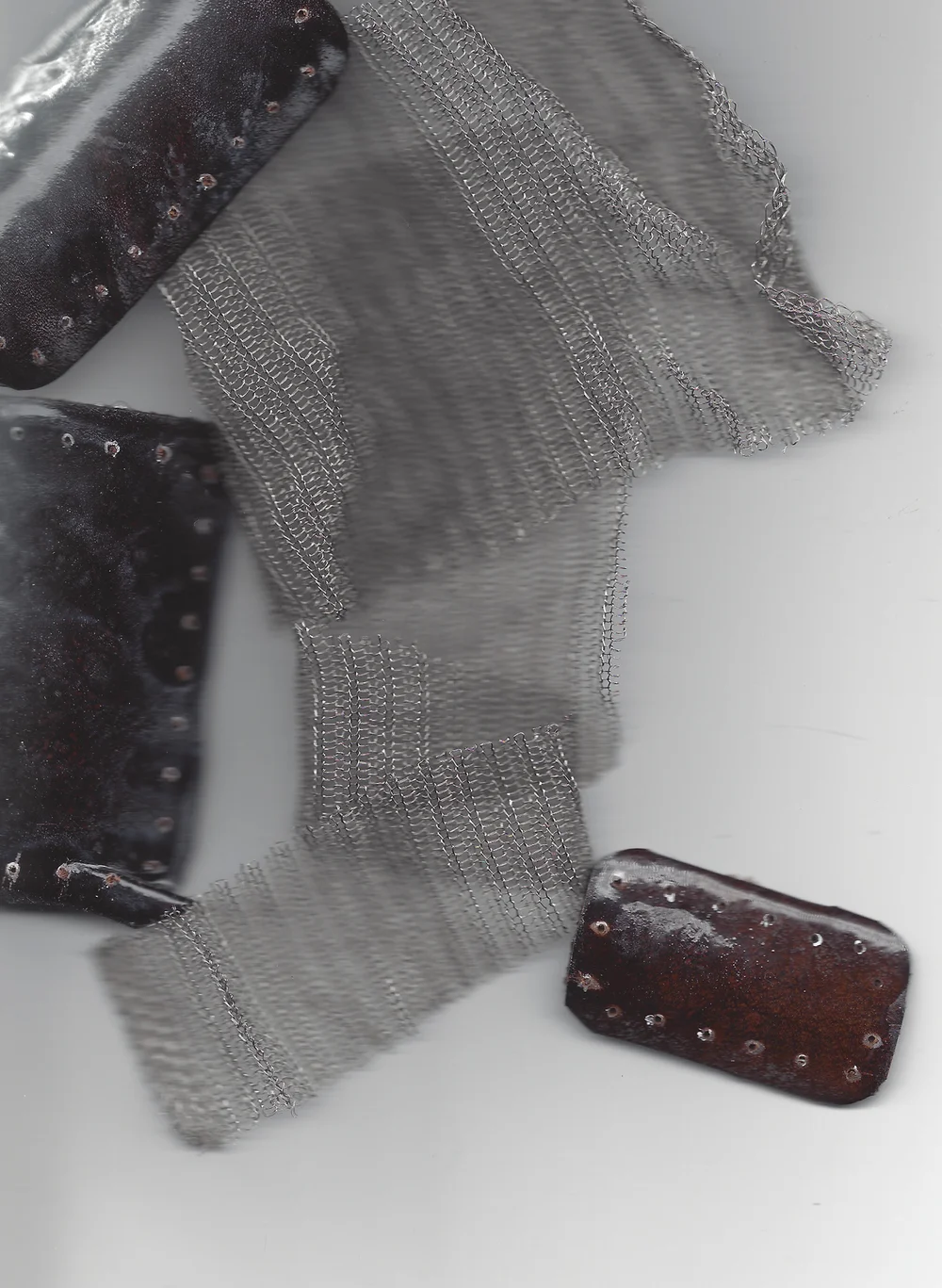 Close-up of dark textured leather-like fabric pieces with metal grommets alongside draped silvery mesh fabric on a neutral background, moody and industrial