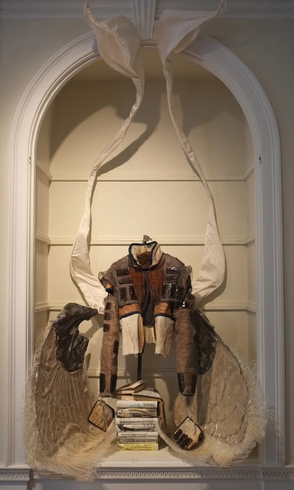 Textured mixed-media garment with wing-like fabric appendages displayed in an arched alcove above a stack of books, neutral gallery setting