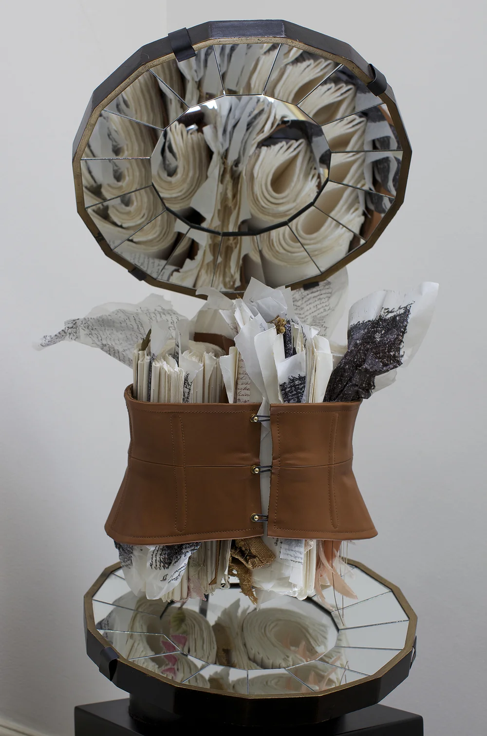 Sculptural art piece combining a mirrored circular frame, layered paper and fabric elements, and a tan leather corset-like form, neutral gallery setting