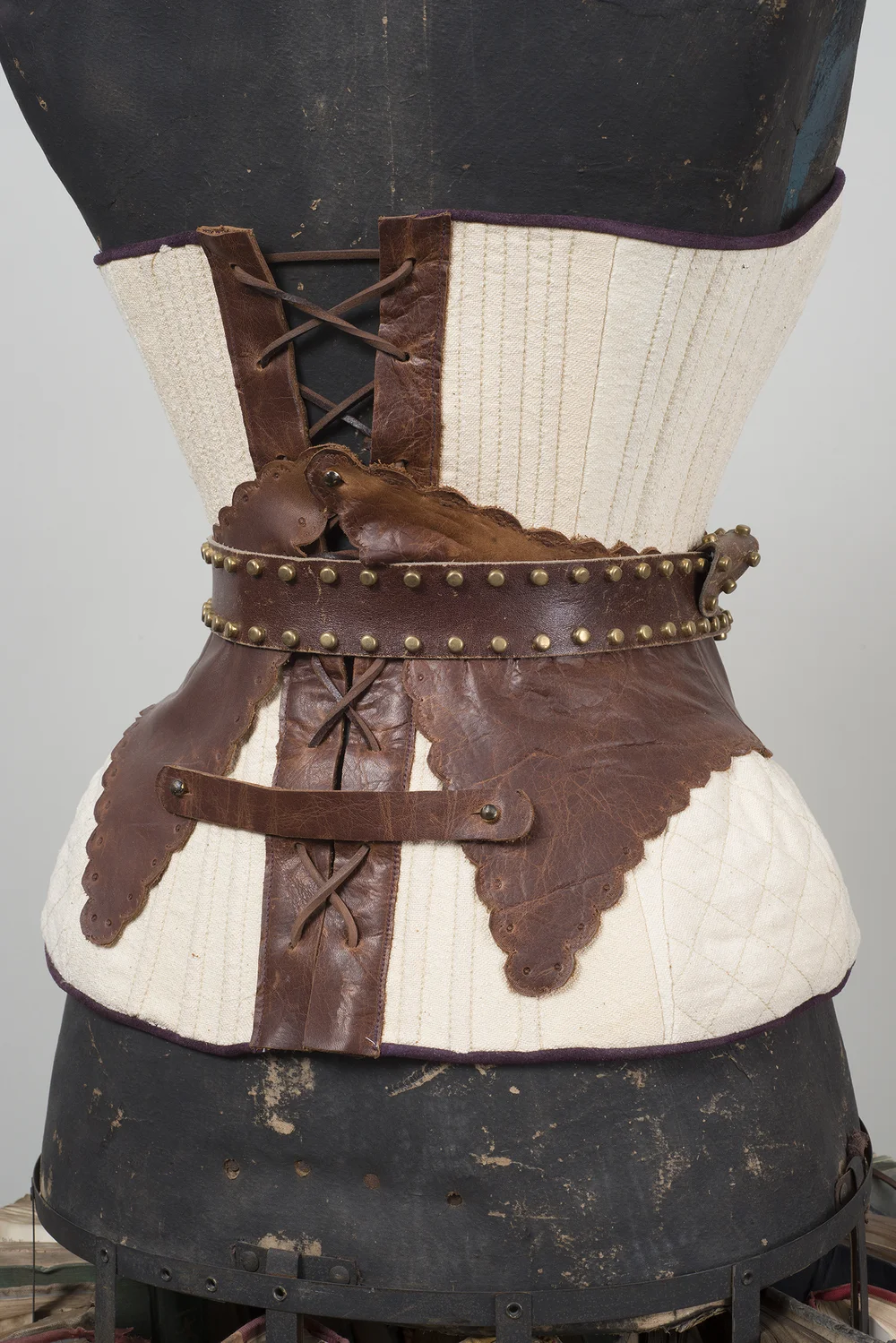 Close-up of a cream and brown leather corset with studded belt and lacing details on a dark dress form, neutral background